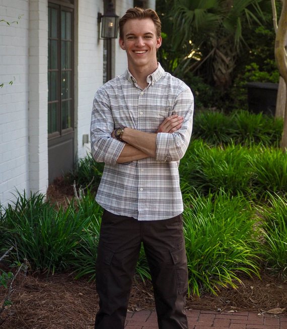 Josiah Falde, arms crossed and smiling, standing on a brick path beside a garden bed